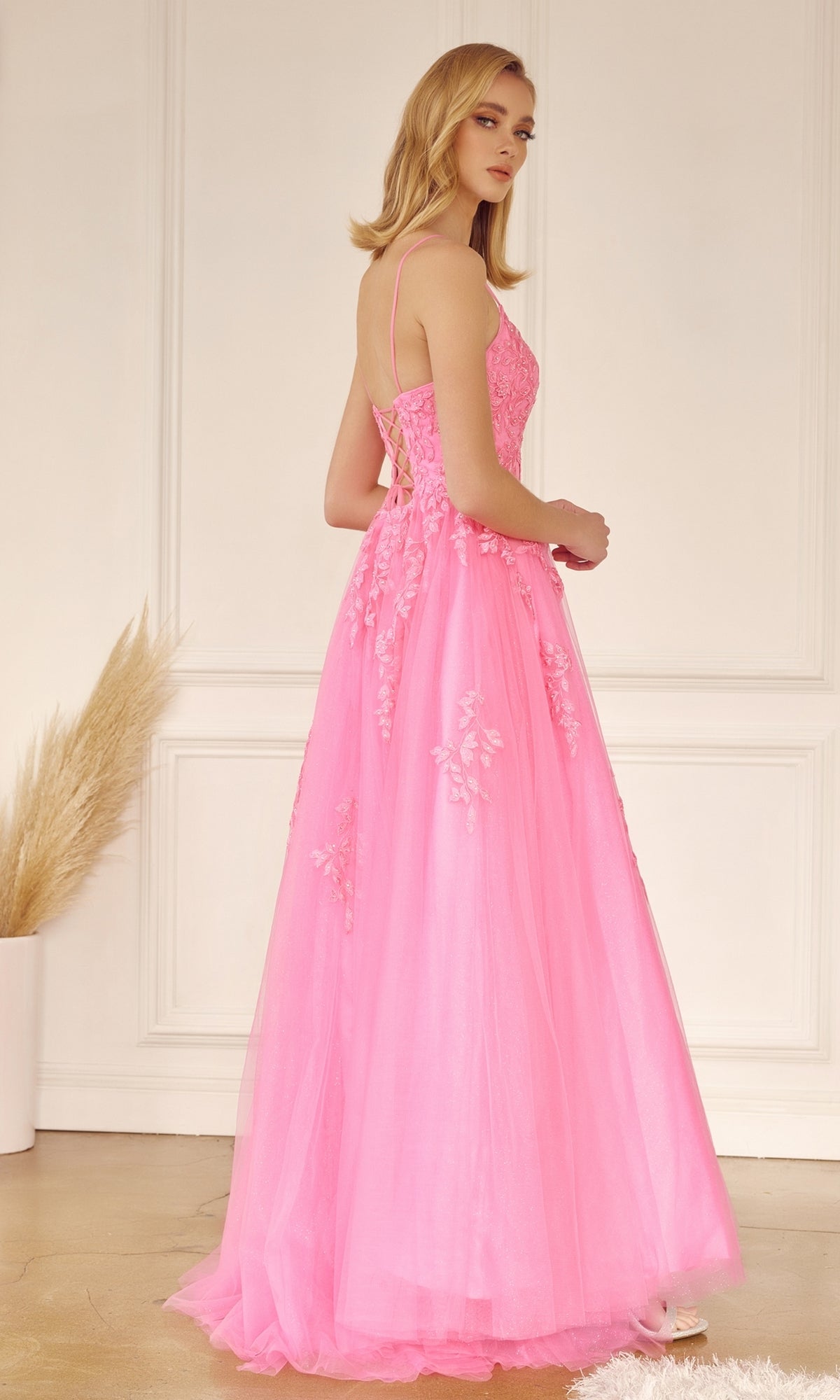 A woman in the Corset-Back Long Glitter-Tulle Prom Ball Gown 260, featuring a floor-length pink tulle design with floral embroidery, stands indoors partially turned from the camera.